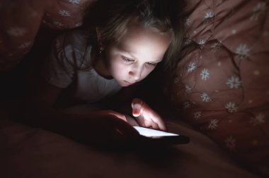 Küçük bir kız uyuduktan sonra dijital tablet akıllı telefon cihazı kullanmak için battaniyenin altına saklanıyor. Küçük çocukların yalnızlığı ve telefonda kurtarma..