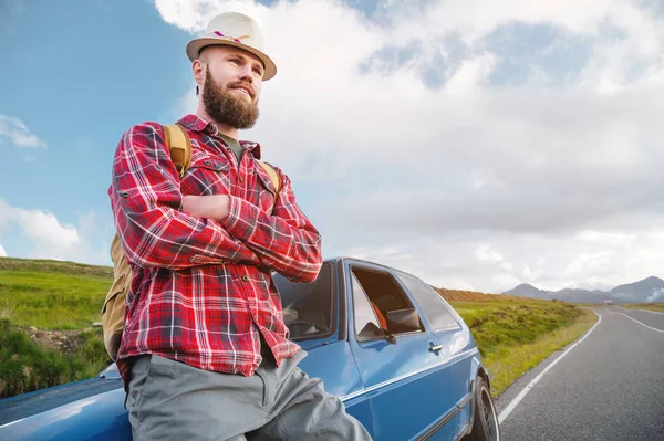 Retrato de um viajante sério feliz barbudo hipster com uma mochila em uma camisa xadrez e um chapéu ao lado de um carro desconhecido fica na estrada ao pôr do sol nas montanhas. Conceito de viagem feliz e confiante — Fotografia de Stock