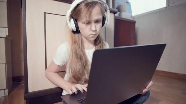 Een slim meisje van zeven met een witte koptelefoon met een laptop in haar handen duwt op de vloer in haar kamer. De jonge generatie op het internet en IT-technologie — Stockfoto