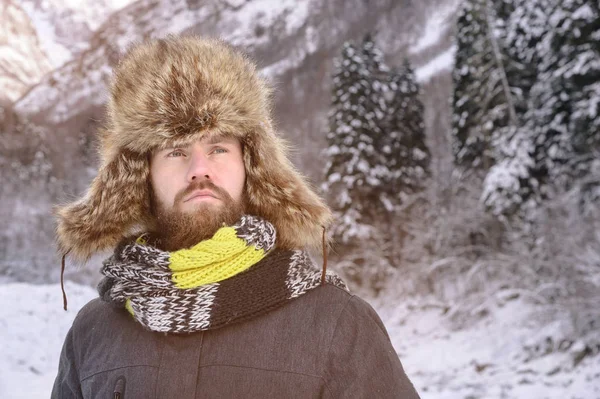 Retrato de um homem viril atraente com bigode e barba em uma floresta de inverno. Turista elegante em um chapéu de pele e um lenço brilhante — Fotografia de Stock