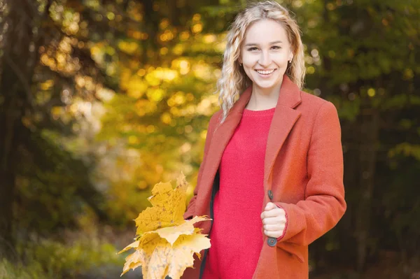 Sonbahar ormanında gülümseyen sarı yapraklarla dolu bir buketle kırmızı paltolu çekici sarışın kızın portresi. Sonbahar, sonbahar tatilleri ve hafta sonları konsepti.