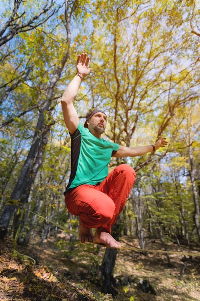 Wide angle male tightrope walker balancing while sitting barefoot on slackline in autumn forest. The concept of outdoor sports and active life of people aged