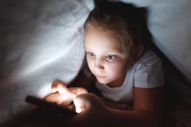 Küçük bir kız uyuduktan sonra dijital tablet akıllı telefon cihazı kullanmak için battaniyenin altına saklanıyor. Küçük çocukların yalnızlığı ve telefonda kurtarma..