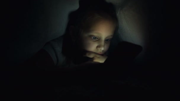 Młoda dziewczynka ukrywa się pod kocem, aby używać cyfrowego tabletu do smartfona późno po zaśnięciu. Samotność małych dzieci i ratunek przez telefon. — Wideo stockowe