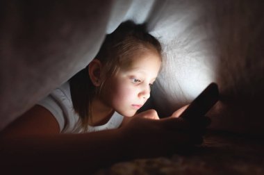 Küçük bir kız uyuduktan sonra dijital tablet akıllı telefon cihazı kullanmak için battaniyenin altına saklanıyor. Küçük çocukların yalnızlığı ve telefonda kurtarma..