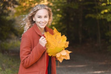 Sonbahar ormanında gülümseyen sarı yapraklarla dolu bir buketle kırmızı paltolu çekici sarışın kızın portresi. Sonbahar, sonbahar tatilleri ve hafta sonları konsepti.