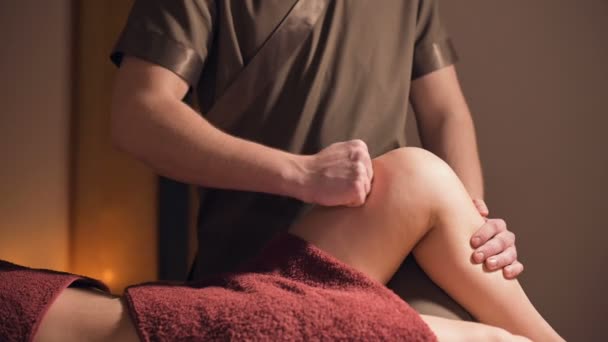 Medium shot of a professional masseur man makes a client massage the legs and knees in an office with dim lighting against the background of burning candles. Premium massage service for luxury clients — Stock Video