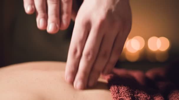 Young male massage therapist is doing finger massage of a woman back with a tattoo in a massage room with dim light on the background of candles. Low key premium massage concept — Stock Video