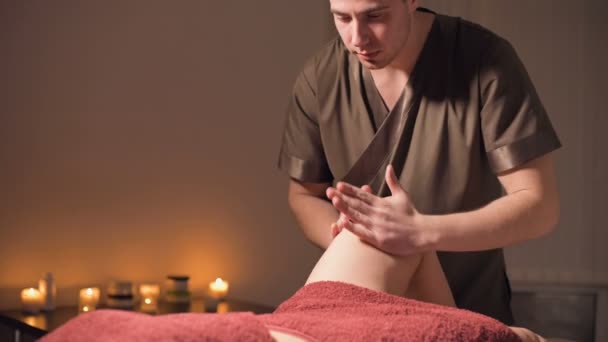 Medium shot van een professionele masseur man laat een klant de benen en knieën masseren in een kantoor met schemerige verlichting tegen de achtergrond van brandende kaarsen. Premium massage service voor luxe klanten — Stockvideo