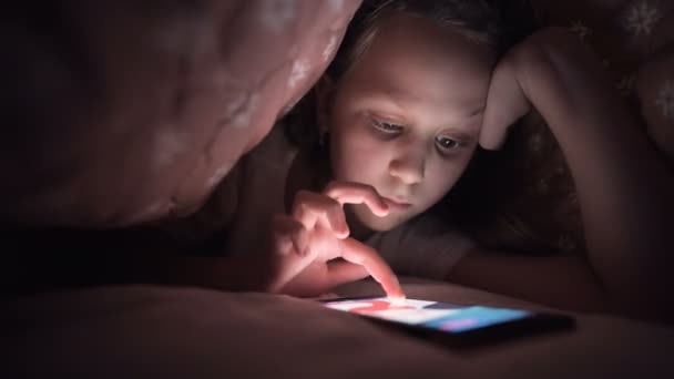 En ung liten flicka gömmer sig under en filt för att använda en digital surfplatta smartphone enhet sent efter sömnen. Ensamhet för små barn och räddning på telefon. — Stockvideo