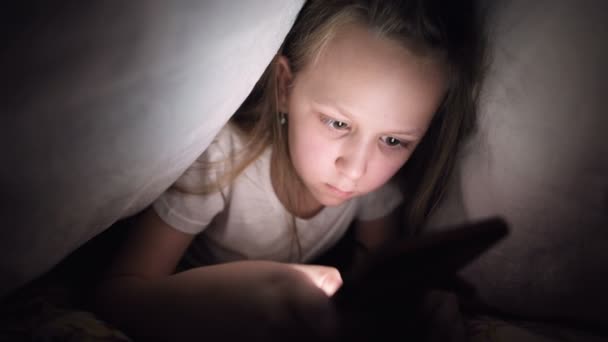 Een jong meisje verstopt zich onder een deken om laat na het slapen een digitaal tablet smartphone apparaat te gebruiken. Eenzaamheid van jonge kinderen en redding aan de telefoon. — Stockvideo