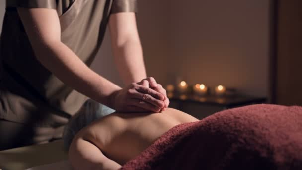 Jonge mannelijke massage therapeut doet rug massage aan een vrouw met een tatoeage in een massageruimte met schemerige lichten op de achtergrond van kaarsen. Low-key premium massageconcept — Stockvideo