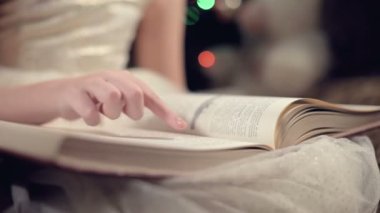Close-up A little blonde girl in a festive dress with a book in her hands sits next to soft toys against the background of a Christmas tree and reads a book leading the page with her place.