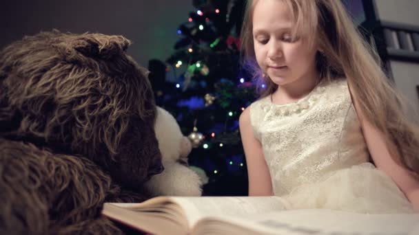 一个穿着节日礼服、手里拿着一本书的金发小女孩坐在一棵圣诞树前的软软玩具旁边，一边看书，一边带着书页. — 图库视频影像