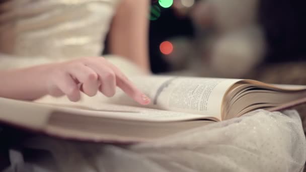 Крупный план Маленькая блондинка в праздничном платье с книгой в руках сидит рядом с мягкими игрушками на фоне елки и читает книгу, ведущую страницу со своим местом . — стоковое видео