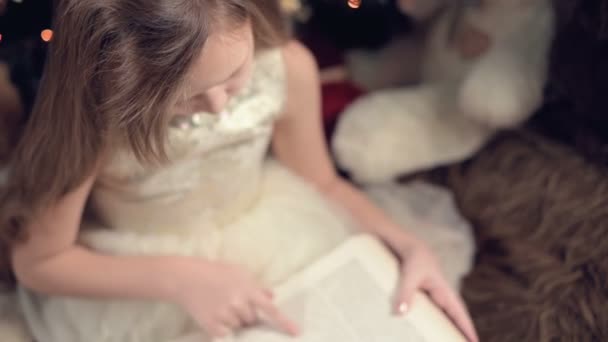Malá blondýnka v svátečních šatech s knihou v ruce sedí vedle měkkých hraček na pozadí vánočního stromečku a čte knihu vedoucí stránku se svým místem. — Stock video
