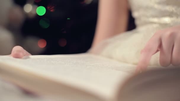 Close-up Een klein blond meisje in een feestelijke jurk met een boek in haar handen zit naast zacht speelgoed tegen de achtergrond van een kerstboom en leest een boek leidt de pagina met haar plaats. — Stockvideo