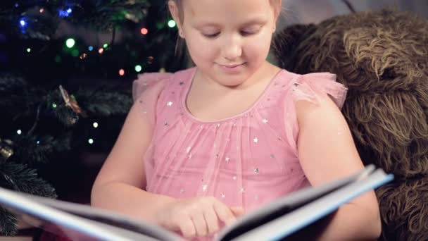 Una bambina bionda in abito festivo con un libro in mano si siede accanto a giocattoli morbidi sullo sfondo di un albero di Natale e legge un libro che conduce la pagina con il suo posto.. — Video Stock
