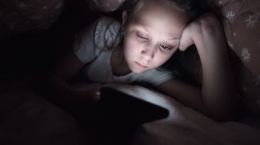 Küçük bir kız uyuduktan sonra dijital tablet akıllı telefon cihazı kullanmak için battaniyenin altına saklanıyor. Küçük çocukların yalnızlığı ve telefonda kurtarma..