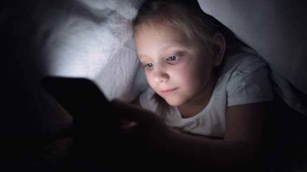 En ung liten flicka gömmer sig under en filt för att använda en digital surfplatta smartphone enhet sent efter sömnen. Ensamhet för små barn och räddning på telefon. — Stockvideo