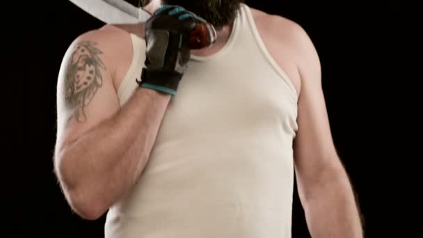Πρυμναίος άντρας με γενειάδα, τατουάζ σε μπλουζάκι σε μαύρο στούντιο που κρατάει μπαλτά. Κλείσιμο — Αρχείο Βίντεο