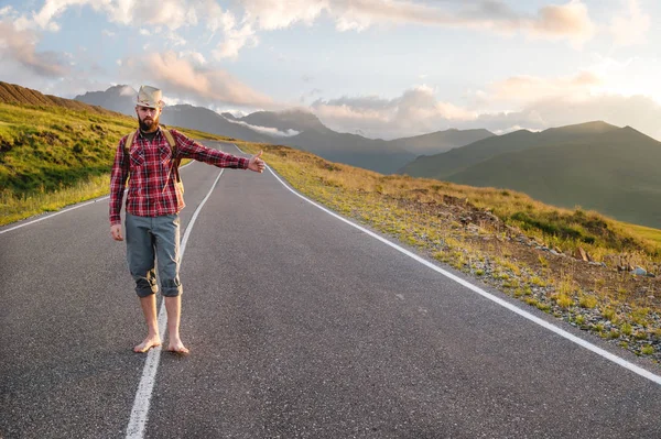 Stílusos mezítlábas szakállas férfi stoppos utazó kalapban és hátizsákkal sétál végig egy külvárosi aszfaltozott úton a hegyekben naplementekor, és megállítja az autót egy stoppal — Stock Fotó