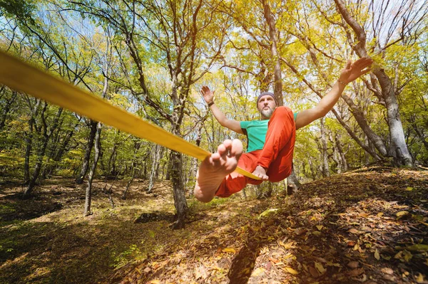 Marcheur de corde serrée mâle grand angle équilibrage tout en étant assis pieds nus sur slackline dans la forêt d'automne. Le concept des sports de plein air et de la vie active des personnes âgées — Photo