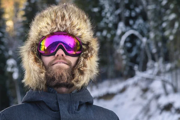 Ritratto ravvicinato di uno sciatore di snowboarder felice barbuto in un passamontagna con occhiali e una pelliccia grande cappello old-school su uno sfondo di montagne innevate invernali — Foto Stock
