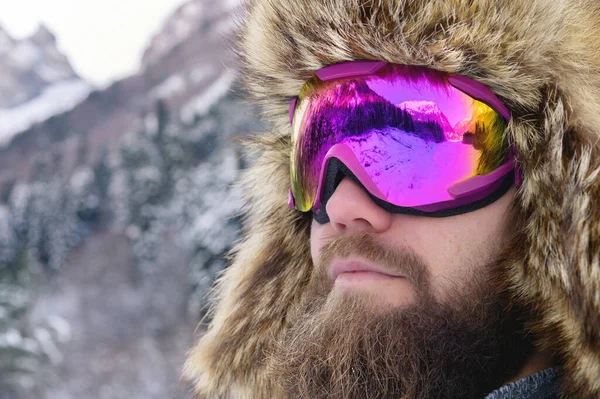 Zbliżenie portret brodatego szczęśliwego narciarza snowboardzisty w masce narciarskiej z goglami i futrzanym starym kapeluszem na tle zimowych gór śnieżnych — Zdjęcie stockowe