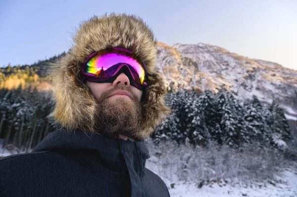 Retrato fechado de um esquiador de snowboard feliz barbudo em uma máscara de esqui com óculos e um chapéu de velha escola grande de pele em um contexto de umas montanhas nevadas de inverno — Fotografia de Stock