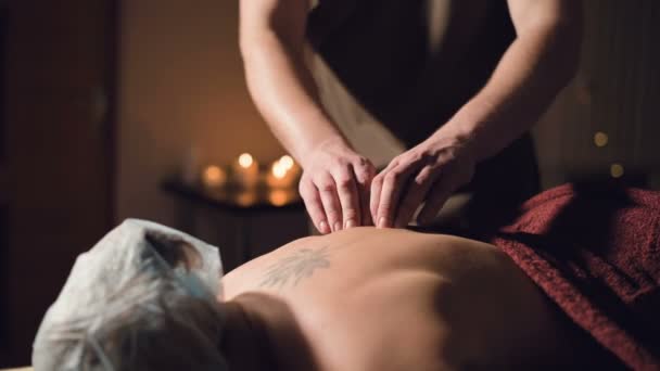 Ung manlig massage terapeut gör rygg massage till en kvinna med en tatuering i ett massagerum med svagt ljus på bakgrunden av ljus. Låg nyckel premium massage koncept — Stockvideo