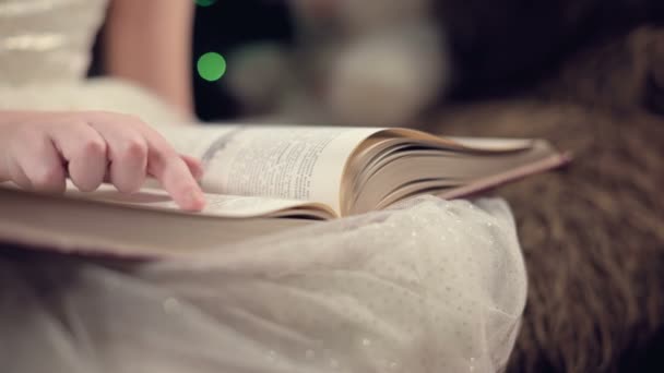 Close-up A little blonde girl in a festive dress with a book in her hands sits next to soft toys against the background of a Christmas tree and reads a book leading the page with her place. — Stok video