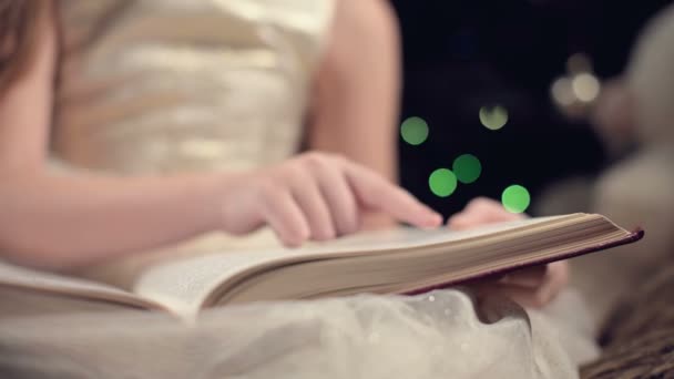 Крупный план Маленькая блондинка в праздничном платье с книгой в руках сидит рядом с мягкими игрушками на фоне елки и читает книгу, ведущую страницу со своим местом . — стоковое видео