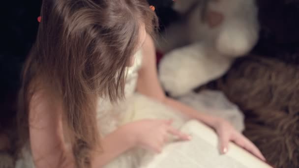 Una niña rubia en un vestido festivo con un libro en las manos se sienta junto a juguetes suaves sobre el fondo de un árbol de Navidad y lee un libro que lleva la página con su lugar. — Vídeos de Stock