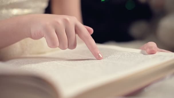 Close-up Una niña rubia en un vestido festivo con un libro en las manos se sienta junto a los juguetes blandos sobre el fondo de un árbol de Navidad y lee un libro que lleva la página con su lugar . — Vídeo de stock