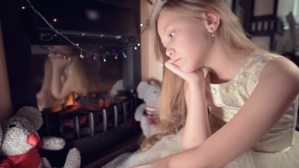 En liten ledsen besviken flicka sitter vid en konstgjord öppen spis och är ledsen bredvid mjuka leksaker. Begreppet bortskämd jullov — Stockvideo