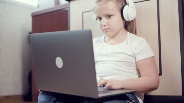 Una niña inteligente de siete años con auriculares blancos con un portátil en las manos está empujando al suelo en su habitación. La generación joven en Internet y la tecnología informática — Vídeos de Stock