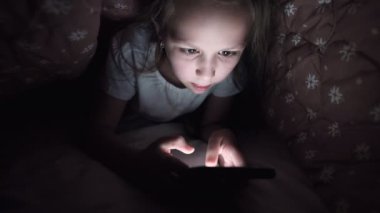 Küçük bir kız uyuduktan sonra dijital tablet akıllı telefon cihazı kullanmak için battaniyenin altına saklanıyor. Küçük çocukların yalnızlığı ve telefonda kurtarma..