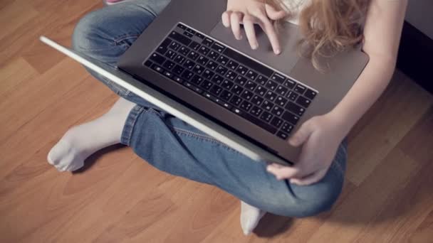 Una niña inteligente de siete años con auriculares blancos con un portátil en las manos está empujando al suelo en su habitación. La generación joven en Internet y la tecnología informática — Vídeo de stock