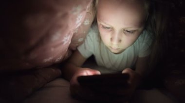 Küçük bir kız uyuduktan sonra dijital tablet akıllı telefon cihazı kullanmak için battaniyenin altına saklanıyor. Küçük çocukların yalnızlığı ve telefonda kurtarma..