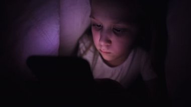 Küçük bir kız uyuduktan sonra dijital tablet akıllı telefon cihazı kullanmak için battaniyenin altına saklanıyor. Küçük çocukların yalnızlığı ve telefonda kurtarma..