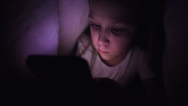 一个小女孩正躲在毯子下，在睡眠不足的时候用数字平板智能手机装置。 年幼儿童的孤独和电话救援. — 图库视频影像