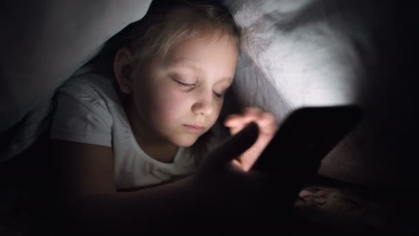 Una niña pequeña se esconde debajo de una manta para usar un dispositivo de teléfono inteligente tableta digital tarde después de dormir. Soledad de los niños pequeños y rescate por teléfono . — Vídeo de stock