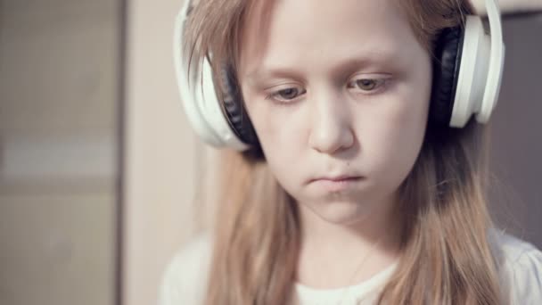 Um close-up de um retrato plano apressado de uma menina séria pensativa e destacada que tem 10 anos de idade em grandes fones de ouvido brancos dentro de casa. Olho lateral — Vídeo de Stock