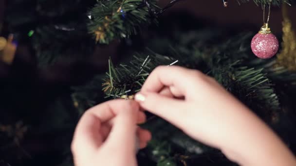 Κοντινό πλάνο του χεριού ενός μικρού κοριτσιού διακοσμεί ένα δέντρο της Πρωτοχρονιάς. Κρεμάει παιχνίδια σε κλαδιά με φόντο γιρλάντες και φώτα — Αρχείο Βίντεο