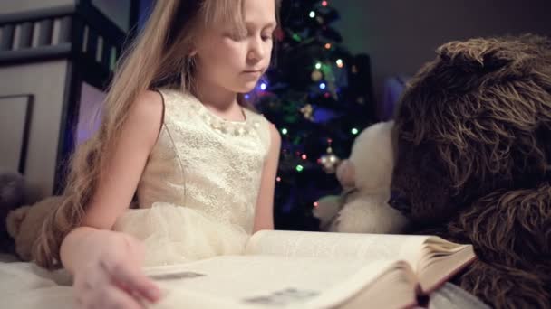 Una niña rubia en un vestido festivo con un libro en las manos se sienta junto a juguetes suaves sobre el fondo de un árbol de Navidad y lee un libro que lleva la página con su lugar. — Vídeo de stock