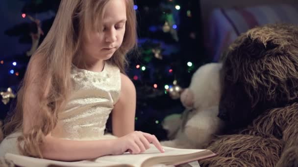 Una niña rubia en un vestido festivo con un libro en las manos se sienta junto a juguetes suaves sobre el fondo de un árbol de Navidad y lee un libro que lleva la página con su lugar. — Vídeos de Stock