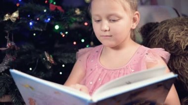 Elinde kitap olan neşeli elbiseli küçük sarışın bir kız Noel ağacının arka planına yaslanmış yumuşak oyuncakların yanına oturur ve kendi evi ile birlikte sayfayı açan bir kitap okur..