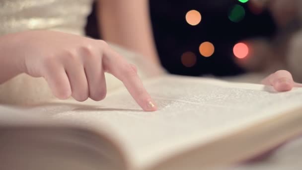 Close-up Mała blondynka w świątecznej sukience z książką w rękach siedzi obok miękkich zabawek na tle choinki i czyta książkę prowadzącą stronę z jej mieszkania. — Wideo stockowe