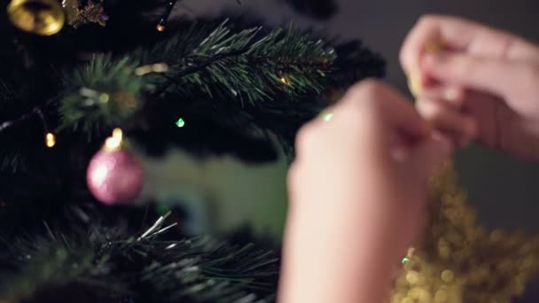 Close-up van de hand van een klein meisje versiert een nieuwjaarsboom. Hangt speelgoed op takken tegen de achtergrond van bloemenslingers en verlichting — Stockvideo